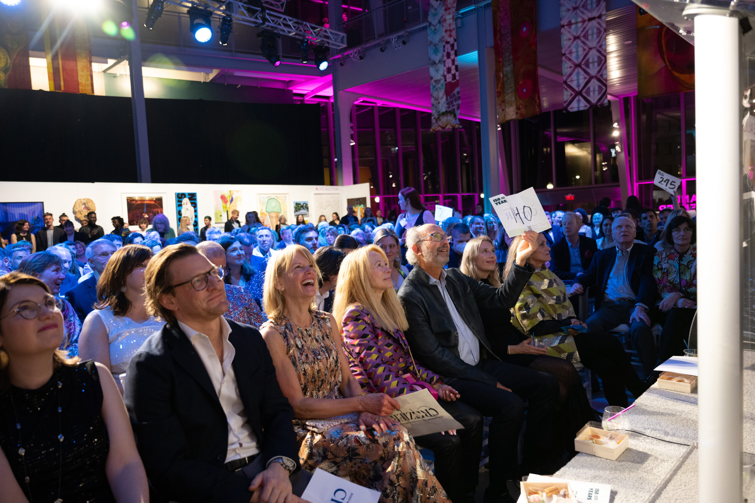 A lively auction scene in a well-lit event space with a seated crowd. Some attendees hold bidding paddles, while others watch attentively. Artworks are displayed in the background, and the atmosphere is vibrant with a mix of colors and lights.