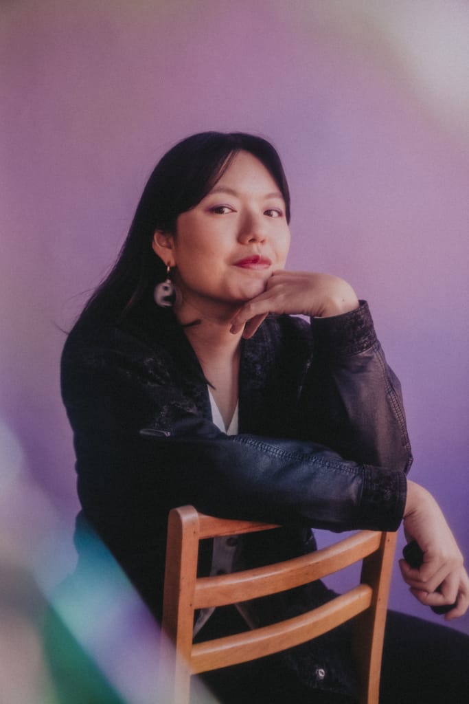 A filmmaker with long dark hair sits on a wooden chair, resting their chin on their hand. They wear a dark jacket and uniquely shaped earrings. The background is a soft blend of purple hues, creating a calm, artistic atmosphere.