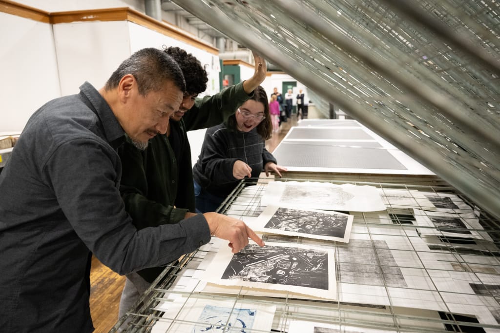 Fred Liang stands with students looking at artwork on a drying rack in the Printmaking studio.