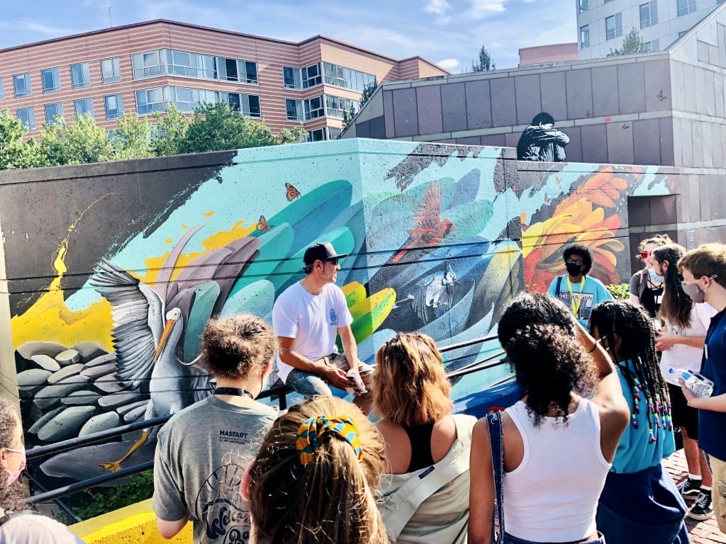 A class of college students stands in front of a teal mural.