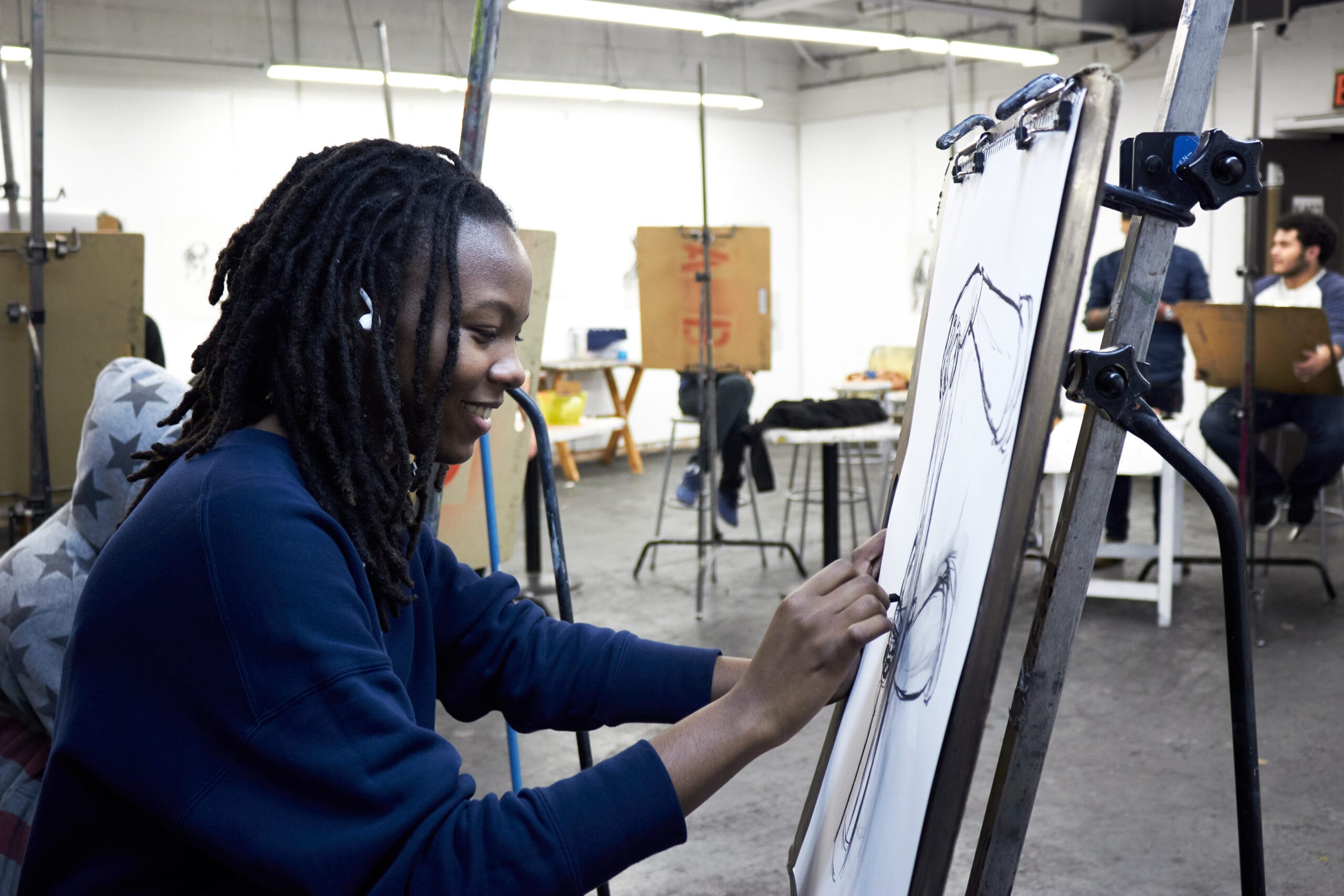 A person with dreadlocks smiles while sketching on a drawing pad in an Artward Bound studio. They wear a dark sweatshirt and earphones. Other artists are visible in the background, each immersed in their own creative journey at their easels.