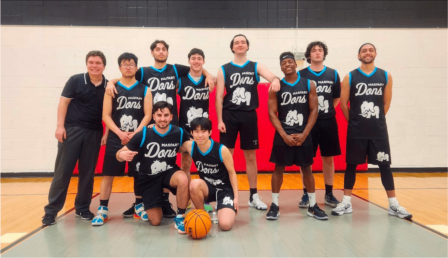 A basketball team poses together in a gym, exuding the spirit of Boston sports. The group includes ten young men, nine in black and teal jerseys and one coach in black athletic wear. One player kneels holding a basketball. The jerseys display an elephant graphic and Dons text, emphasizing their love for recreation.
