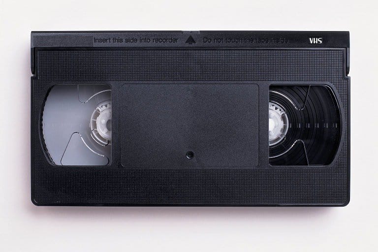 A VHS cassette tape lies on a white surface, featuring a black plastic casing. The tapes clear spools with magnetic tape are visible on either side, with labeling instructions at the top edge for insertion direction.