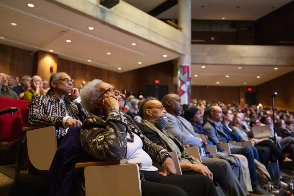 In a large auditorium reminiscent of bustling classrooms, a diverse audience listens attentively, focusing on an elderly woman with glasses in the foreground. The room buzzes with energy, embodying the spirit of a thriving formal event or lecture.