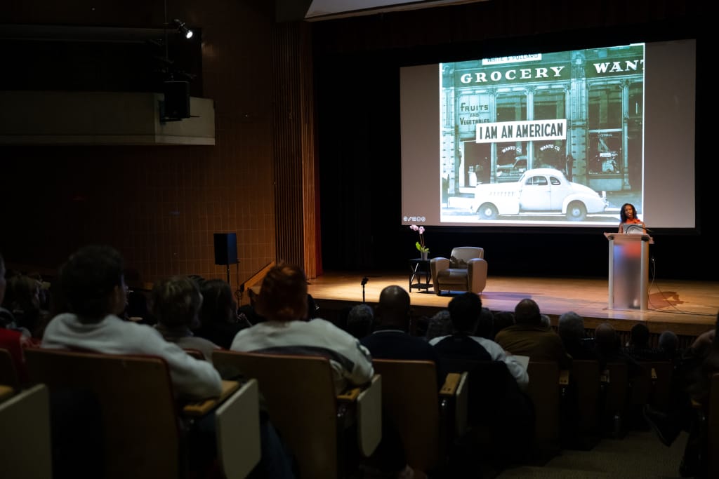 During the Lecture Series, Tyrone Maurice Adderley captivates an audience in a dimly lit auditorium. A historical black-and-white photo of a storefront reading “I Am An American” is projected on the screen behind him, while attendees sit attentively in theater-style rows.