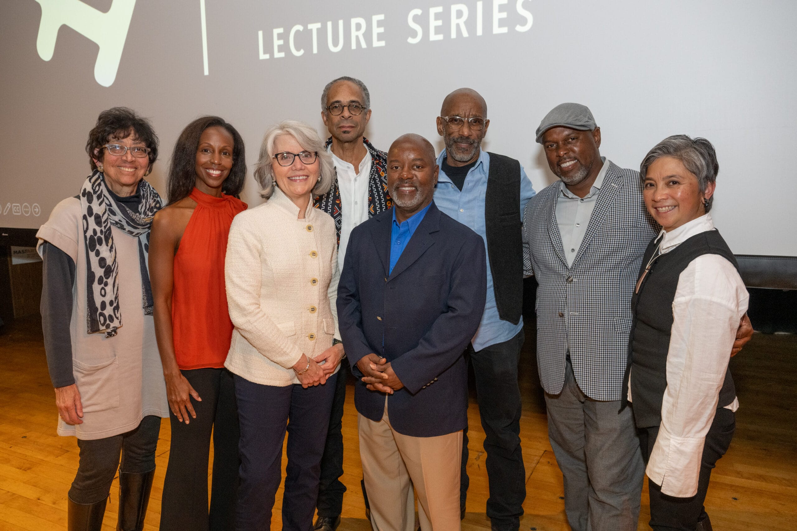 A group of nine people, including Tyrone Maurice Adderley, stands together at the Lecture Series event. They pose in front of a large screen, all smiling and dressed in a stylish mix of casual and formal attire.