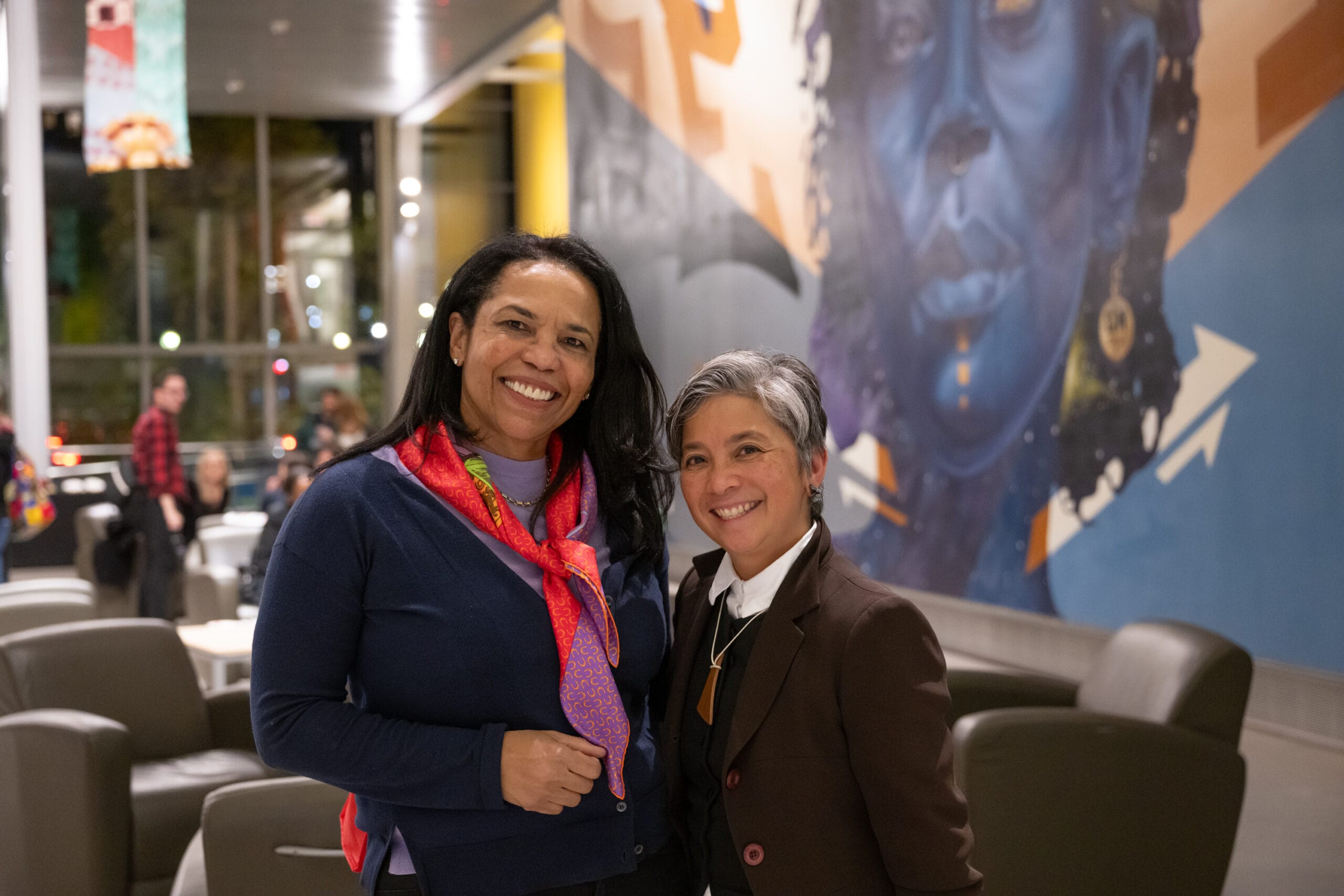 Two people smile in a modern indoor space during the Fall 2023 Tyrone Maurice Adderley Lecture Series. One wears a dark top and pink scarf, the other a brown blazer and white shirt. They stand before an abstract mural, with chairs and attendees in the background, discussing Vision & Justice by Sarah Elizabeth Lewis.