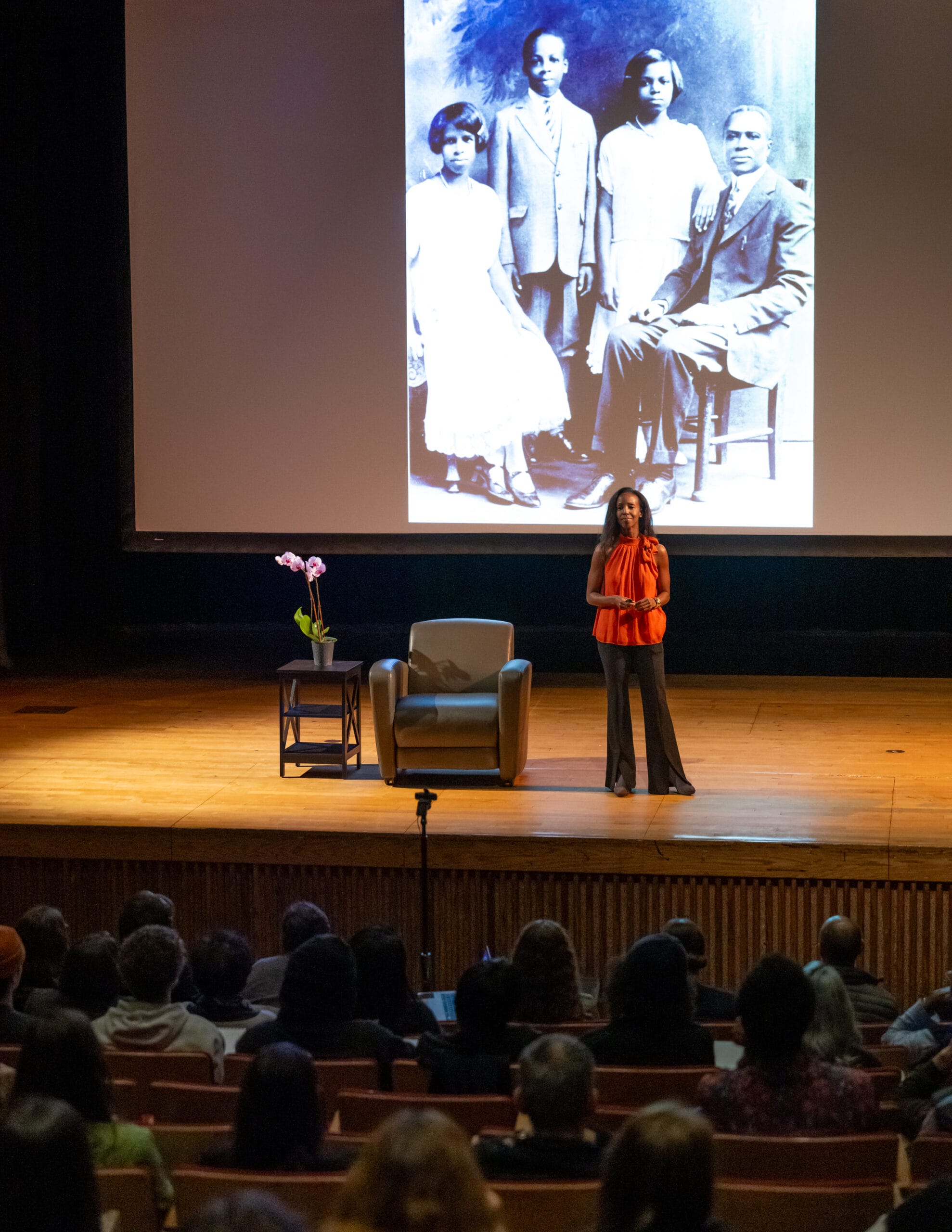 Tyrone Maurice Adderley stands on a stage in front of an attentive audience, with a large historical family photograph projected behind him. The stage features a chair and a table with flowers, setting the tone for his captivating lecture series presentation.