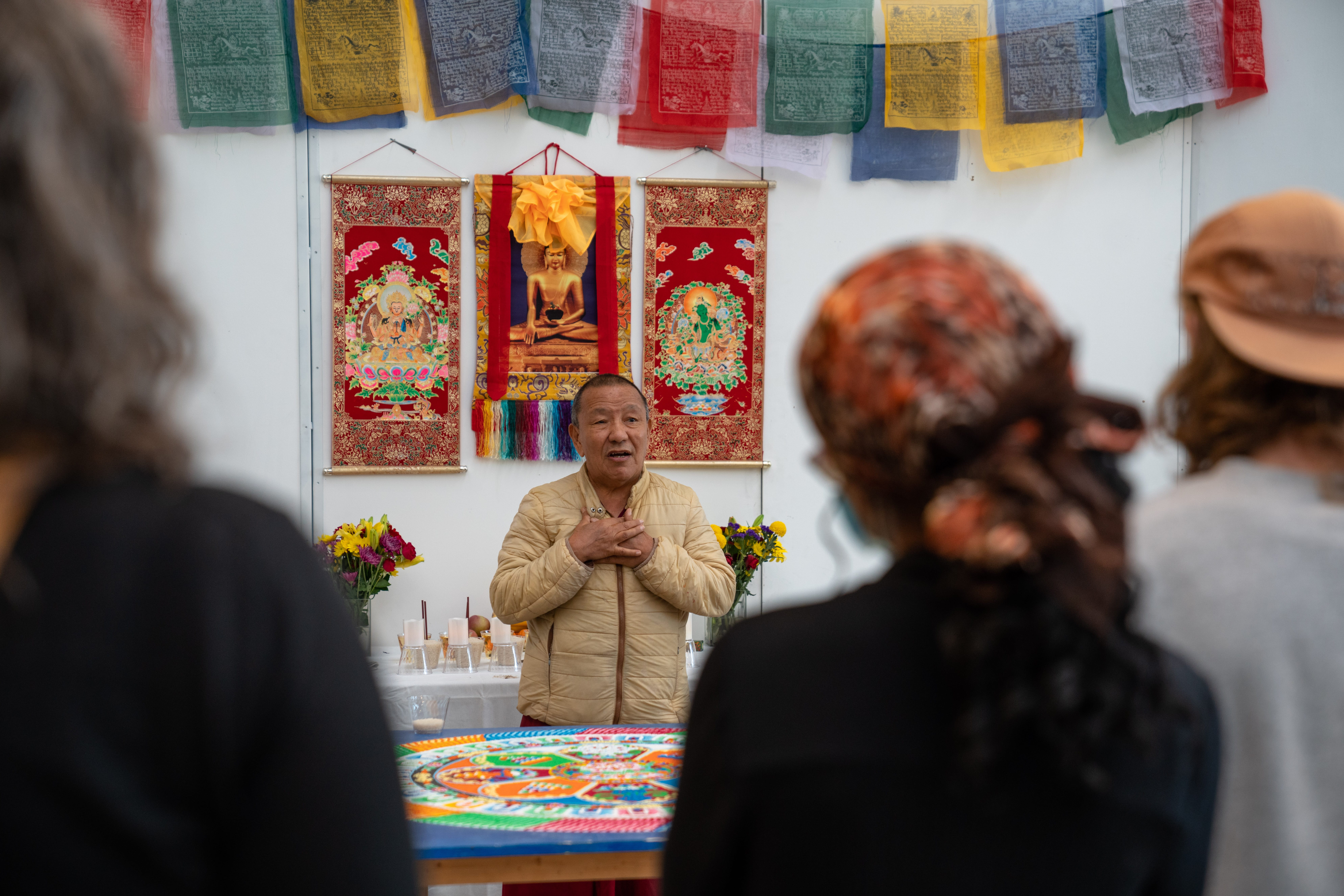 A man stands in front of a sand mandala, speaking to onlookers.