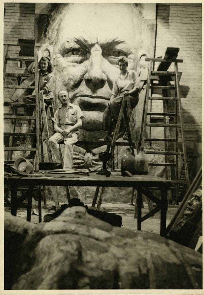 Three people are posing on ladders in front of a large, detailed sculpture of a face mounted on a wall. Art supplies and tools are scattered on tables and the floor in the foreground.