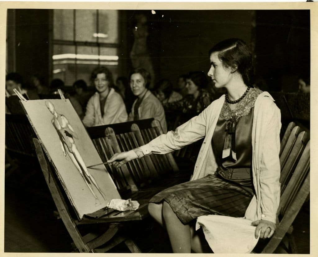 A woman in a blouse and skirt sits on a wooden chair, painting a human figure on an easel. She holds a palette and brush, focused on her work. Several people sit in the background, observing. The setting appears to be a classroom or studio.