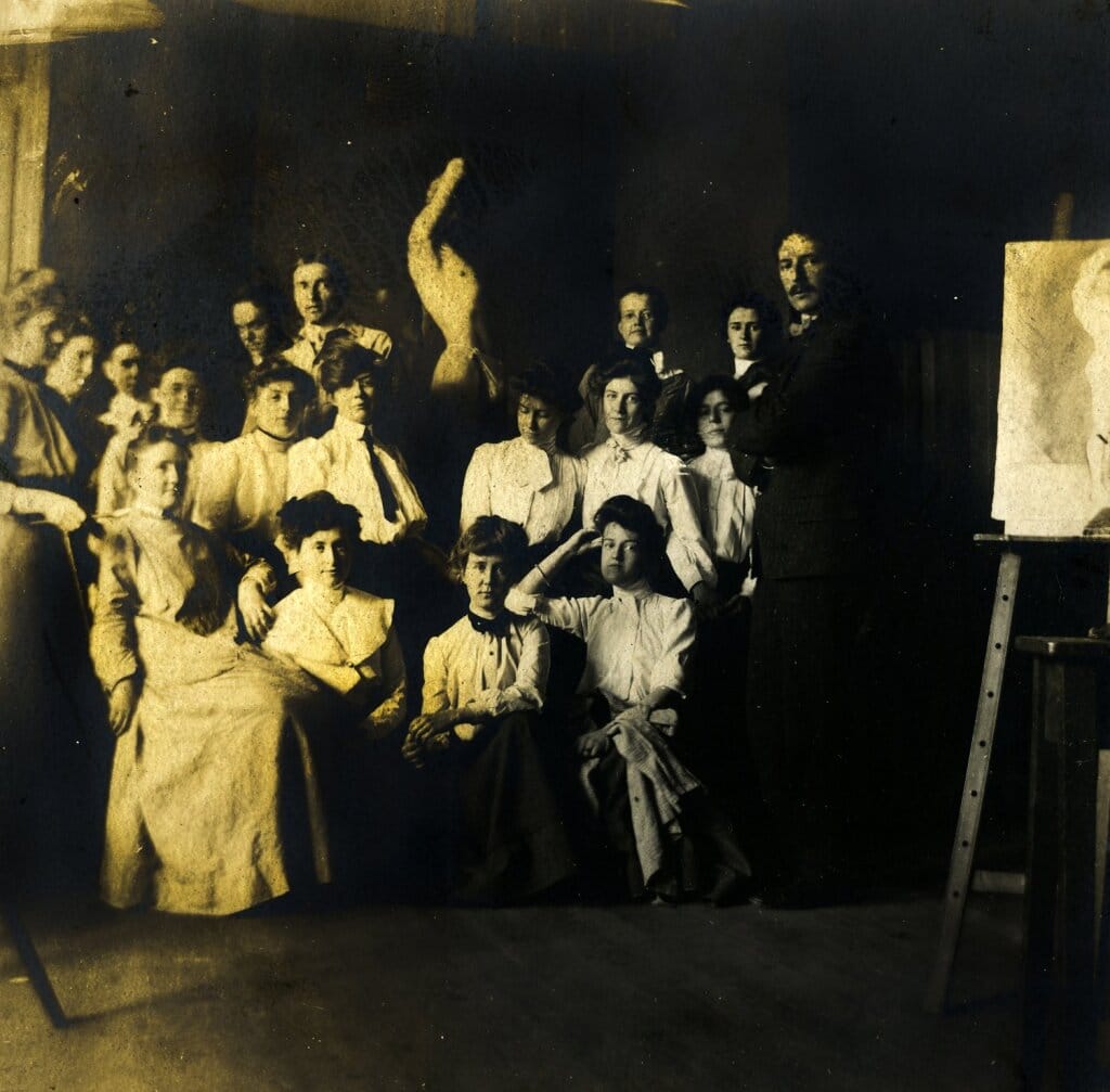 A group of women and a man in vintage clothing are gathered in a dimly lit room. Some are seated, and others stand around a sculpture. An easel with a painting is visible on the right. The image captures an early 20th-century art or gathering.