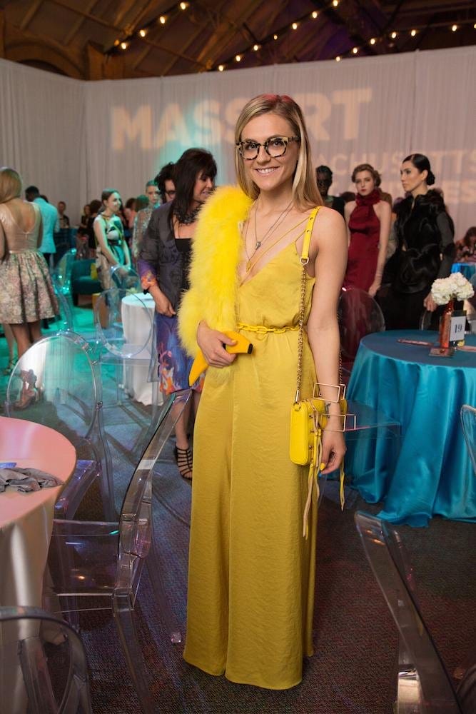 A woman wearing a bright yellow jumpsuit and glasses stands in a stylish indoor event. She has a yellow feathered accent and a matching bag. People are mingling in the background near round tables with blue tablecloths.