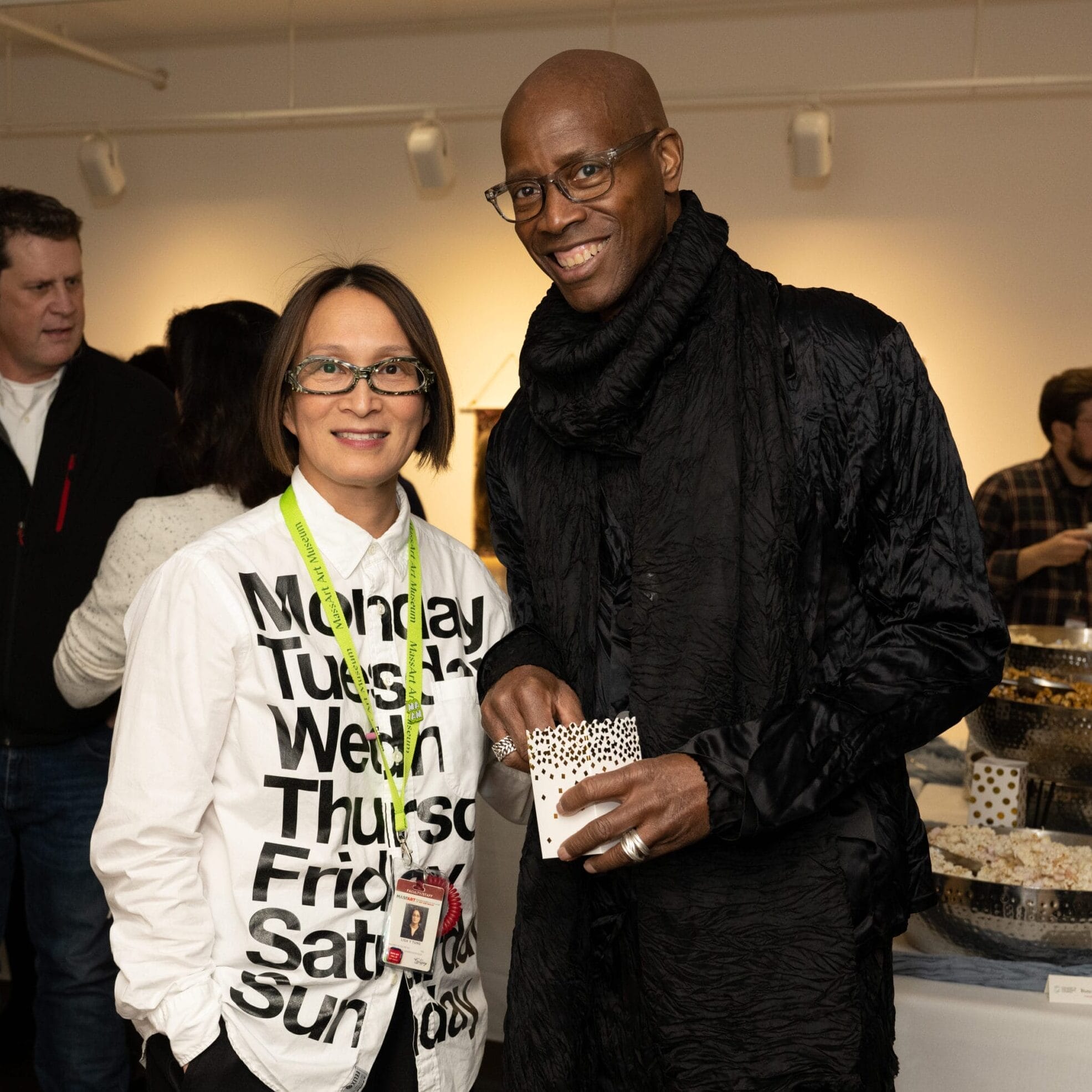 A man in a black outfit and glasses stands next to a woman in a shirt with the days of the week written on it at what appears to be a MassArt gathering. The atmosphere is casual and social, with other people mingling around a table filled with snacks.