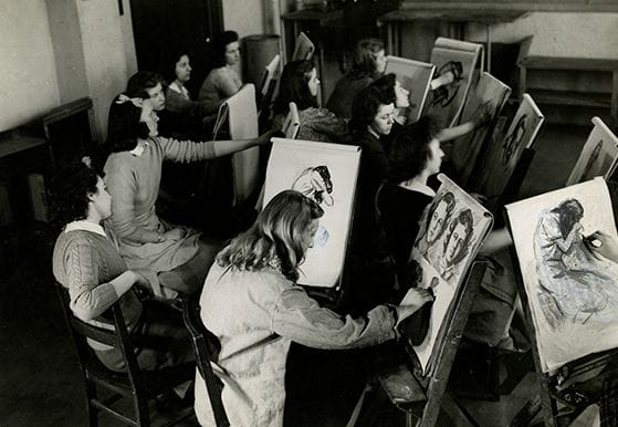 A group of students in a classroom seated at easels, focused on sketching portraits. The room is filled with wooden chairs and artwork in progress. The atmosphere is studious and creative.