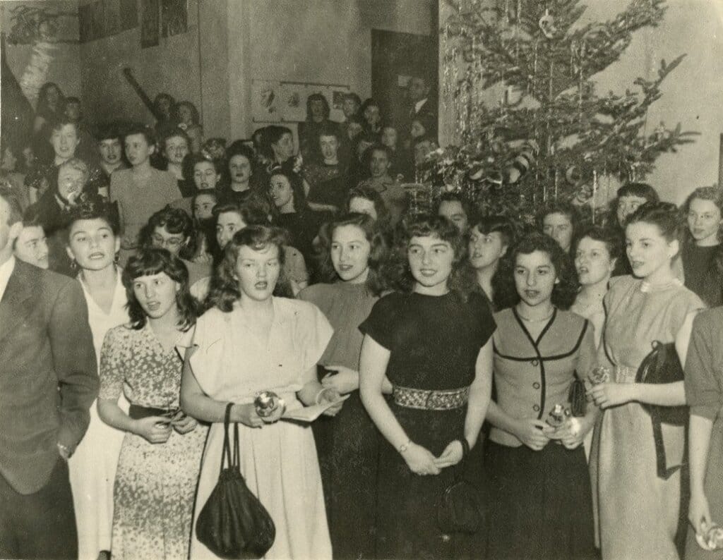 A vintage black-and-white photo shows a large group of people, mostly women, gathered around a decorated Christmas tree. The women are dressed in mid-20th-century attire, and some are holding small gifts. The setting appears festive and lively.
