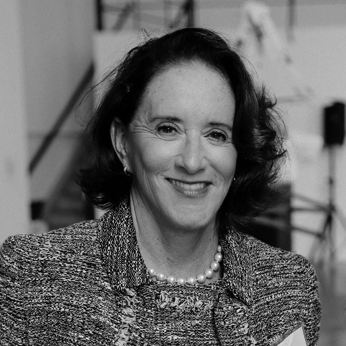 A person with shoulder-length dark hair, wearing a patterned jacket and pearl necklace, is smiling. The background blurs into indistinct shapes and forms. This black-and-white image captures a timeless elegance, as if they belong to a distinguished Board of Trustees.
