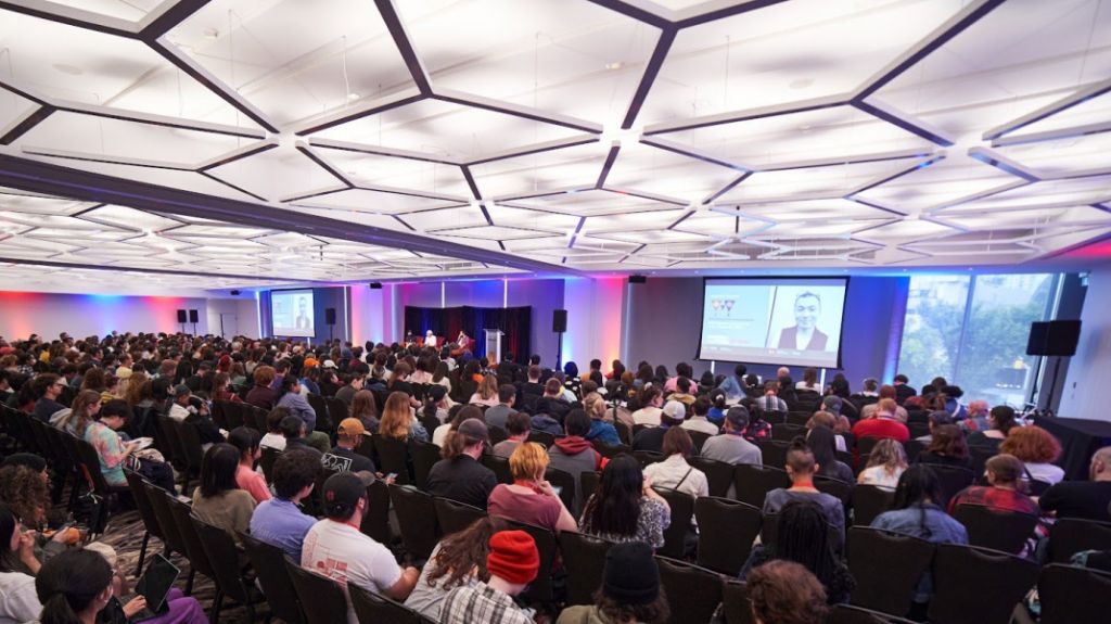 A purple lit room full of a seated audience with a geometric white grid of lights on the ceiling