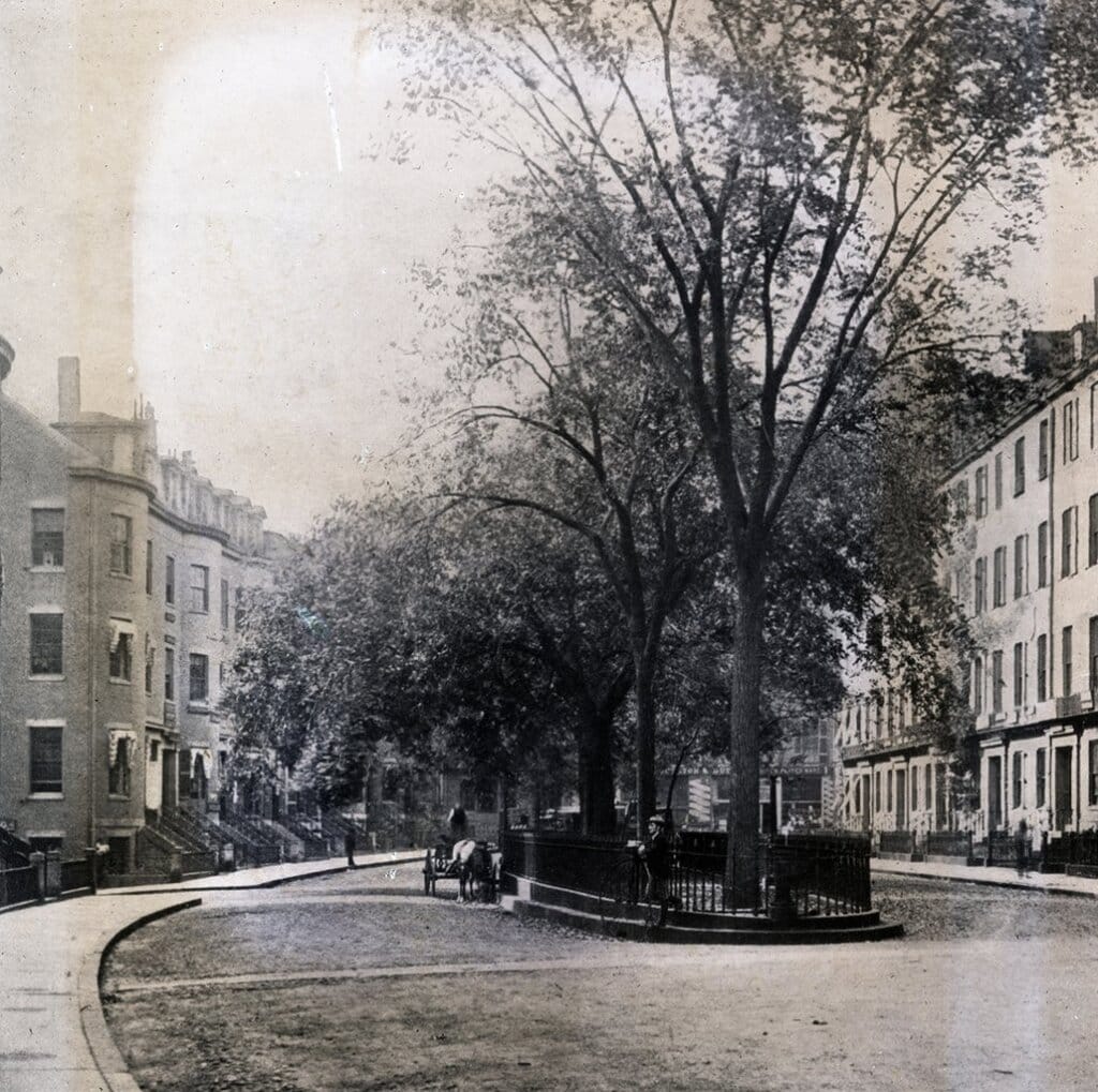 A vintage black-and-white photo of a quiet, curved street lined with multi-story brick row houses. Tall trees stand in the center of the street, and a few people are walking. A horse-drawn cart is visible. The scene is peaceful and historic.