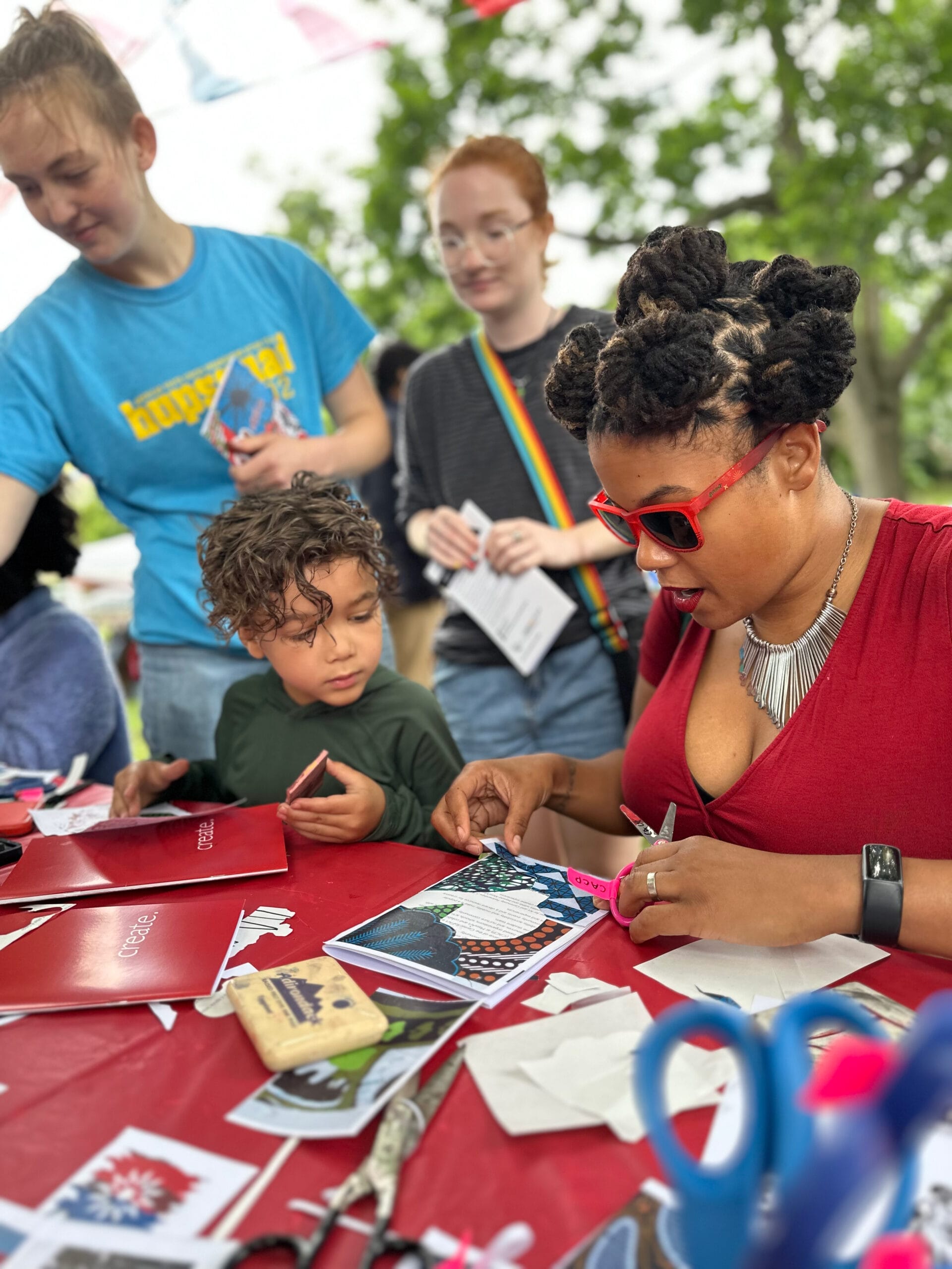 A woman with sunglasses sits at an outdoor table doing a craft with a child.