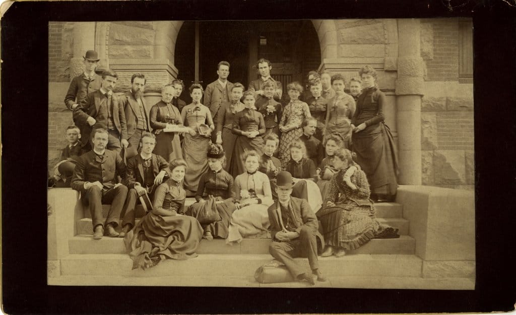A vintage sepia-toned photo shows a group of men and women in late 19th-century attire. They are posing on stone steps in front of a building, with women in dresses and men in suits and hats, creating a formal but amicable atmosphere.