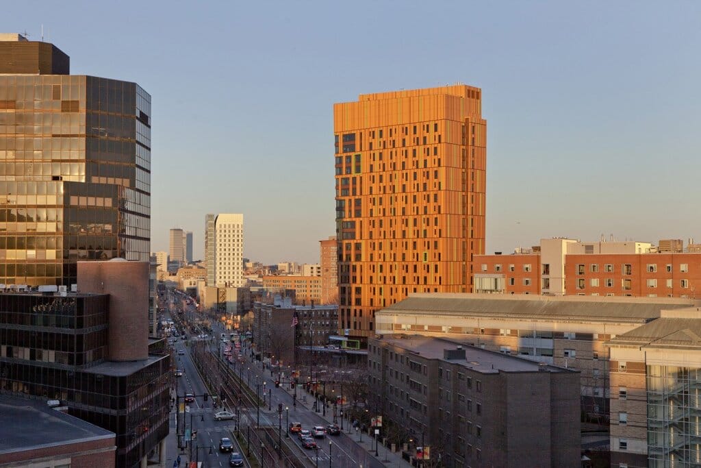 A cityscape view shows a tall, modern orange building contrasting with surrounding structures. A busy street lined with trees and cars runs between them. The sky is clear and the sun casts a warm glow on the scene.