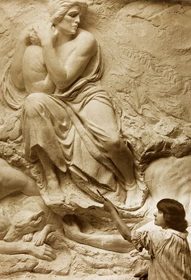 A black and white photo shows a woman working on a large sculpture relief. She holds a tool, refining the details of a draped figure carved into the stone. The sculpture depicts a seated, draped figure above and other shapes below.