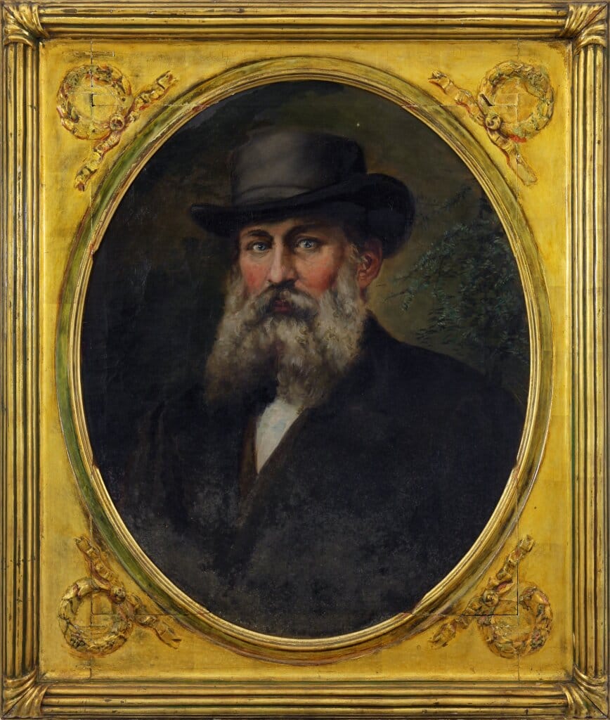 An oval-framed portrait of an older man with a long white beard and a black hat, wearing a dark coat. The frame is ornate and gilded, featuring intricate designs. The background is dark, highlighting the subjects face.