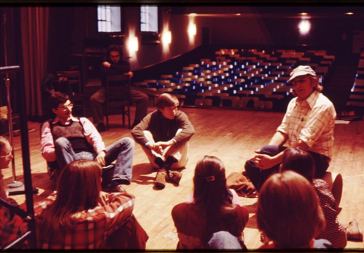 A group of people sit in a circle on a wooden stage. One person in a cap and plaid shirt appears to be leading the discussion. Empty seats are visible in the background. The lighting is warm and dim.