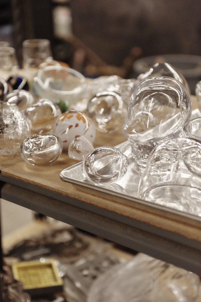 A variety of clear glass objects are displayed on a table, including spheres and abstract shapes. Some objects contain colored elements. The table surface is light-colored, and the background is blurred.