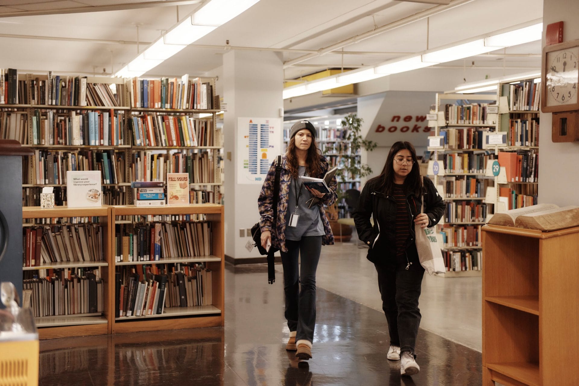 Two people walk through a library. They are surrounded by shelves filled with books. One person is holding a notebook and wearing headphones, while the other carries a bag. A sign in the background reads new books.