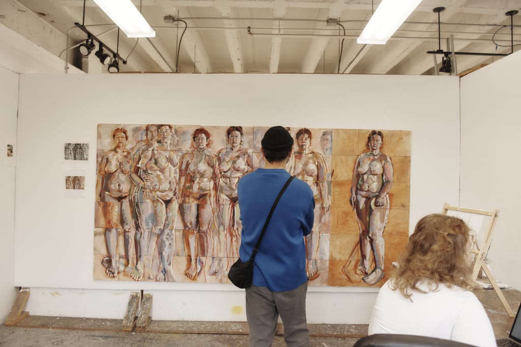 A person in a blue shirt and black hat stands as the image hero in front of a large, detailed painting depicting multiple nude figures in earthy tones. Another person with curly hair sits nearby, partially visible. The room, with its overhead lights, exudes an art studio vibe.