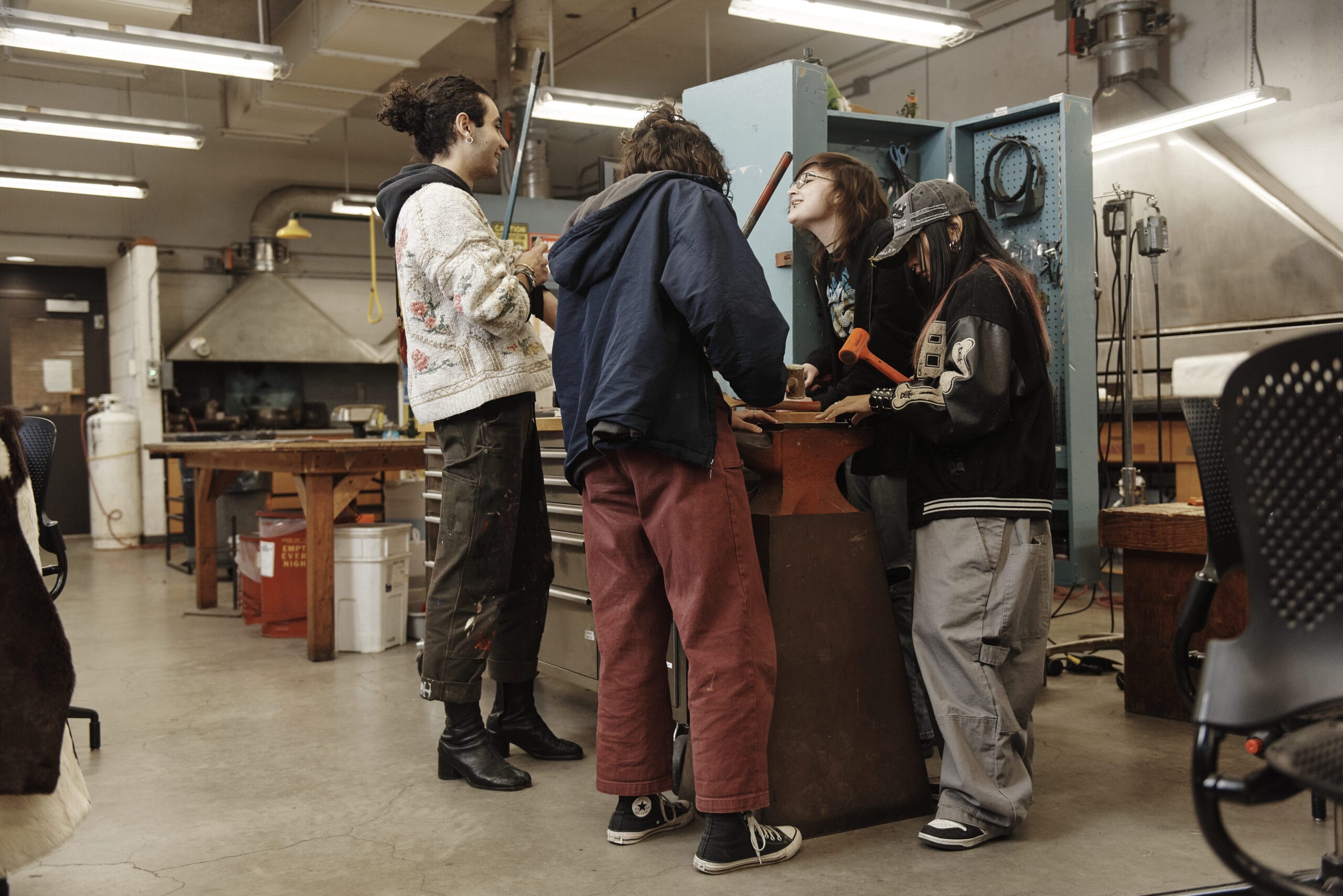 Four people in casual outfits stand around a workbench in a workshop. One is using an orange tool while the others observe. The space is filled with equipment and tools, and they appear engaged in a discussion.