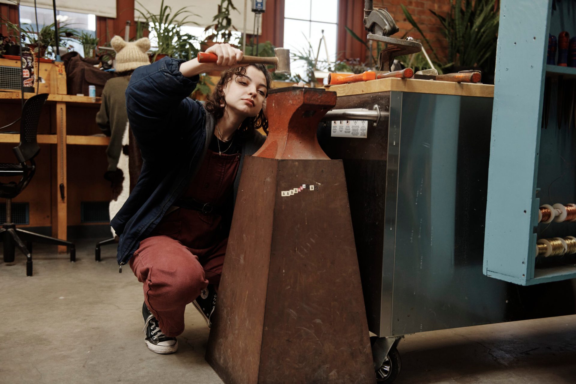 A person is kneeling in a workshop, using a hammer on a metal object. They are wearing a blue jacket and red overalls. The workshop has various tools and equipment in the background.