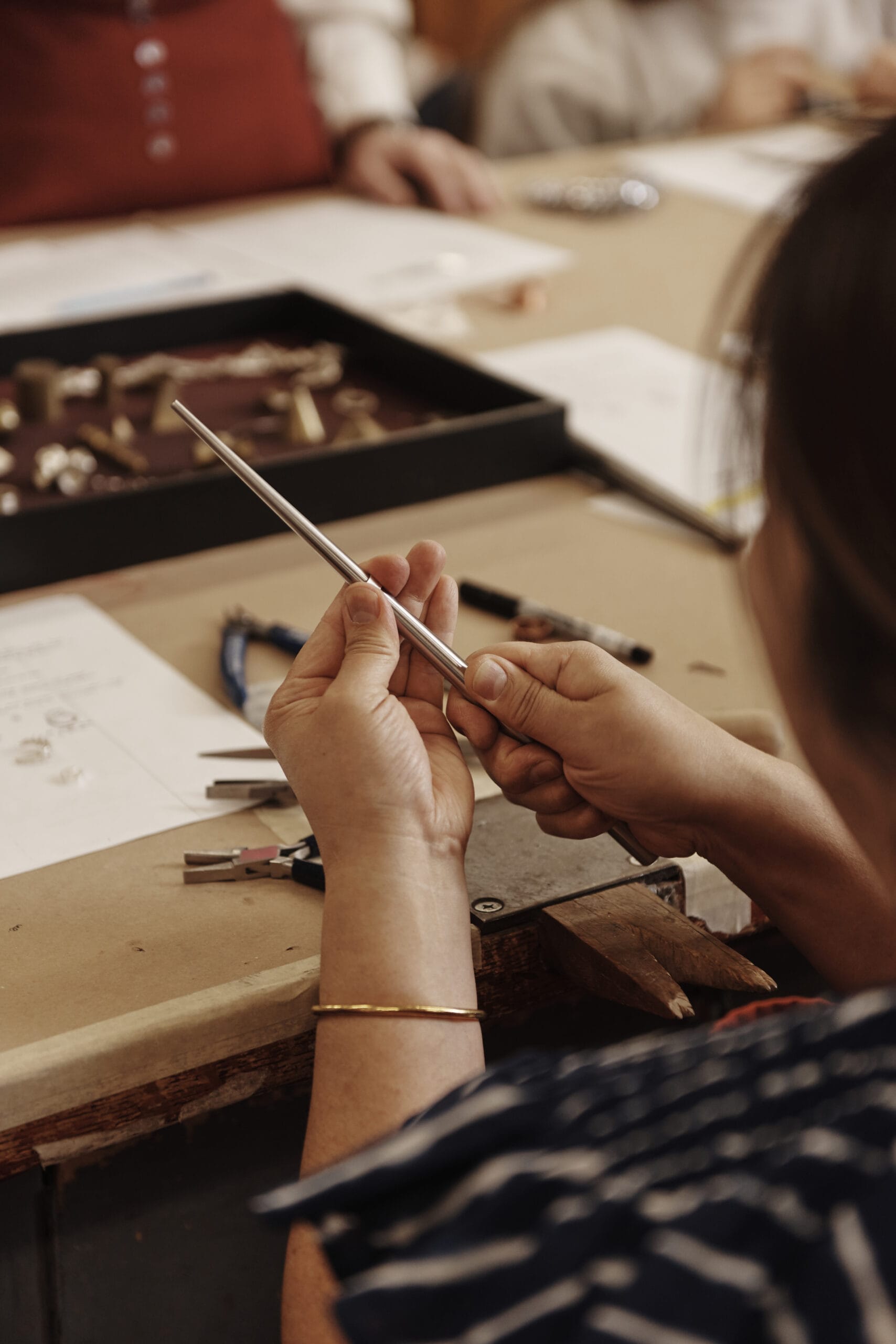 A person holds a metal rod while working at a crafting table. Various tools, papers, and small parts are spread on the table. The person wears a striped shirt and a thin bracelet. Others are sitting nearby, partially visible in the background.