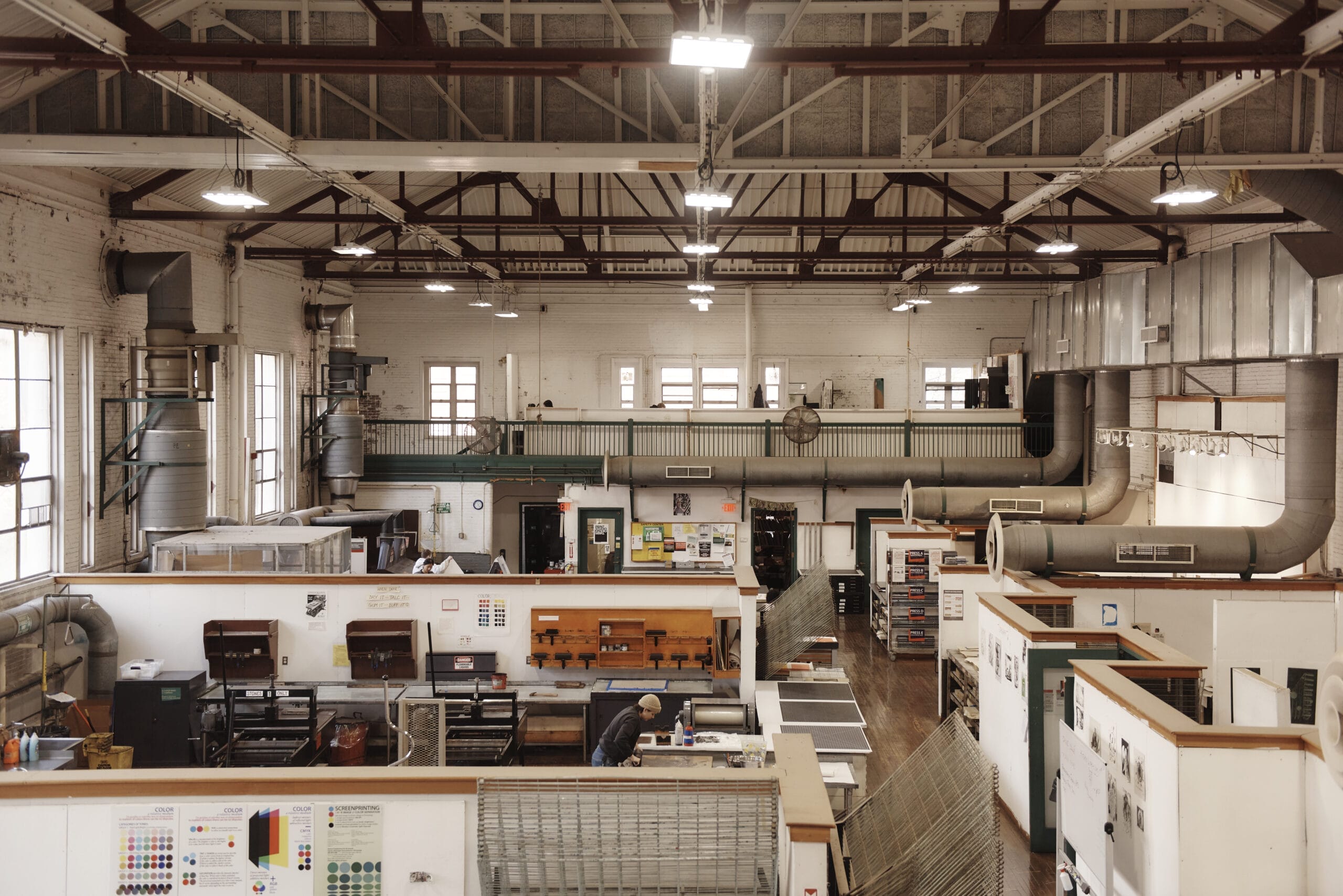 Industrial workspace with high ceilings and large windows, featuring printing equipment and organized workstations. Metal ductwork runs along the ceiling. A person is working at a table, surrounded by shelves and machinery.