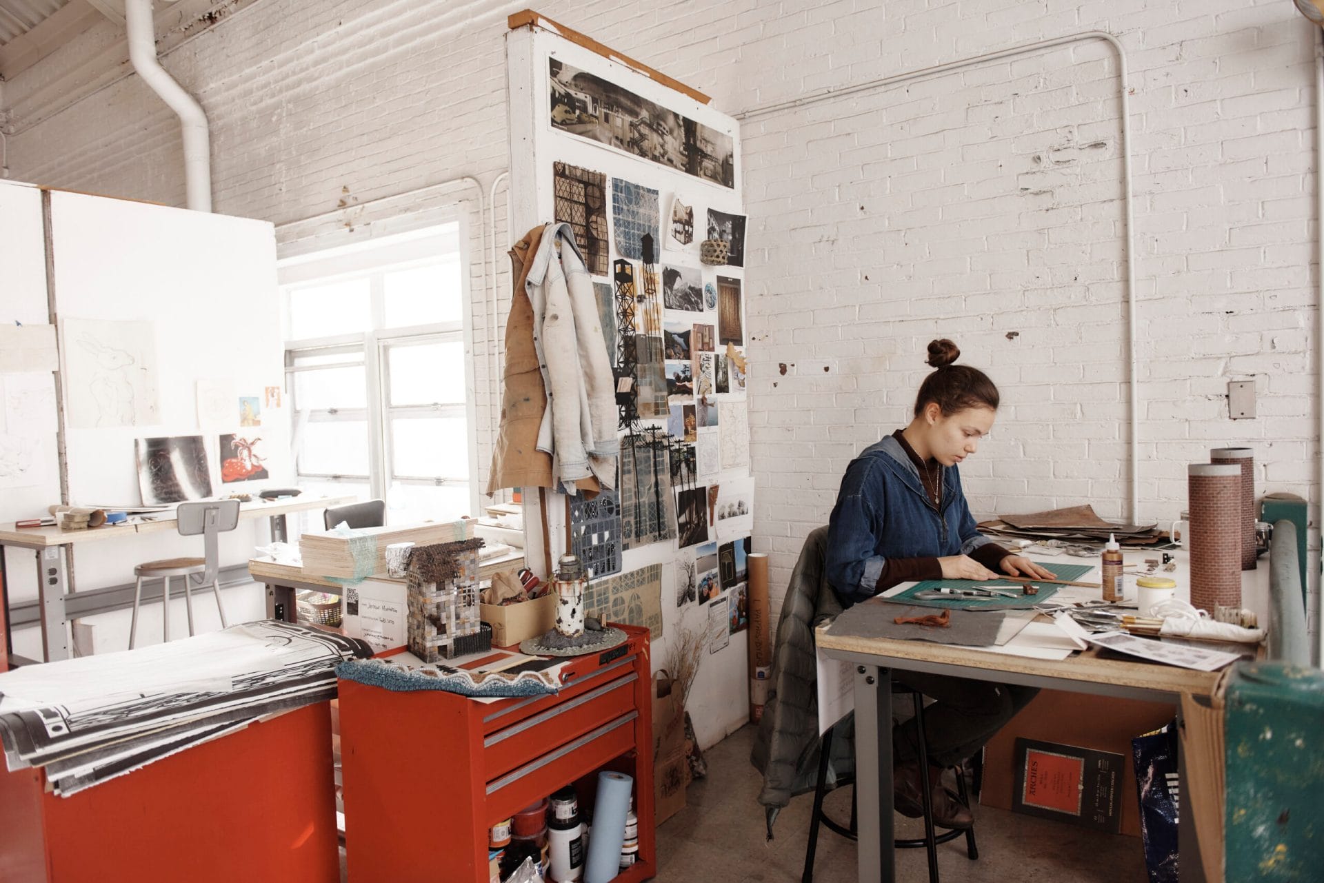 Woman concentrating on a project at a desk in an art studio, surrounded by art supplies and inspiration images on a wall. Shes wearing a denim shirt and has her hair in a bun. The room has a white brick wall and natural light from windows.