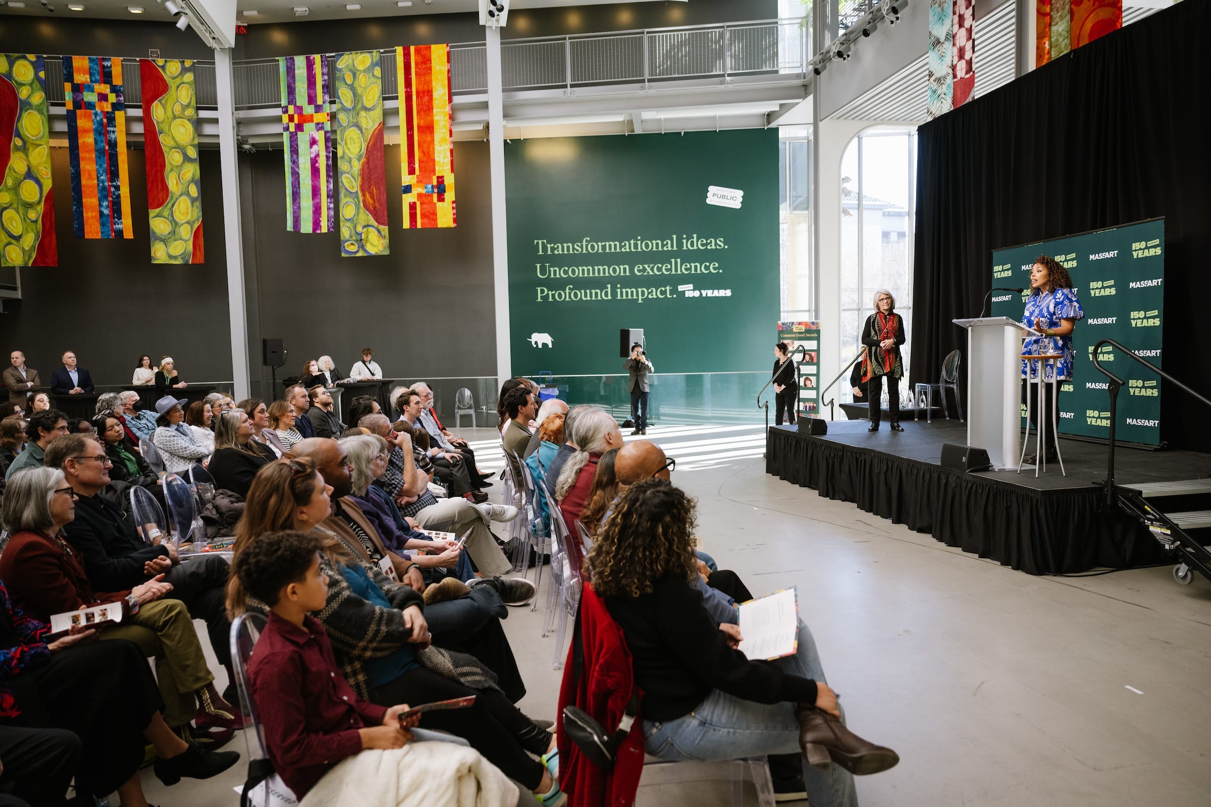 A woman stands at a pedestal on a stage while talking to an audience full of people in MassArt's Design and Media Center Atrium.