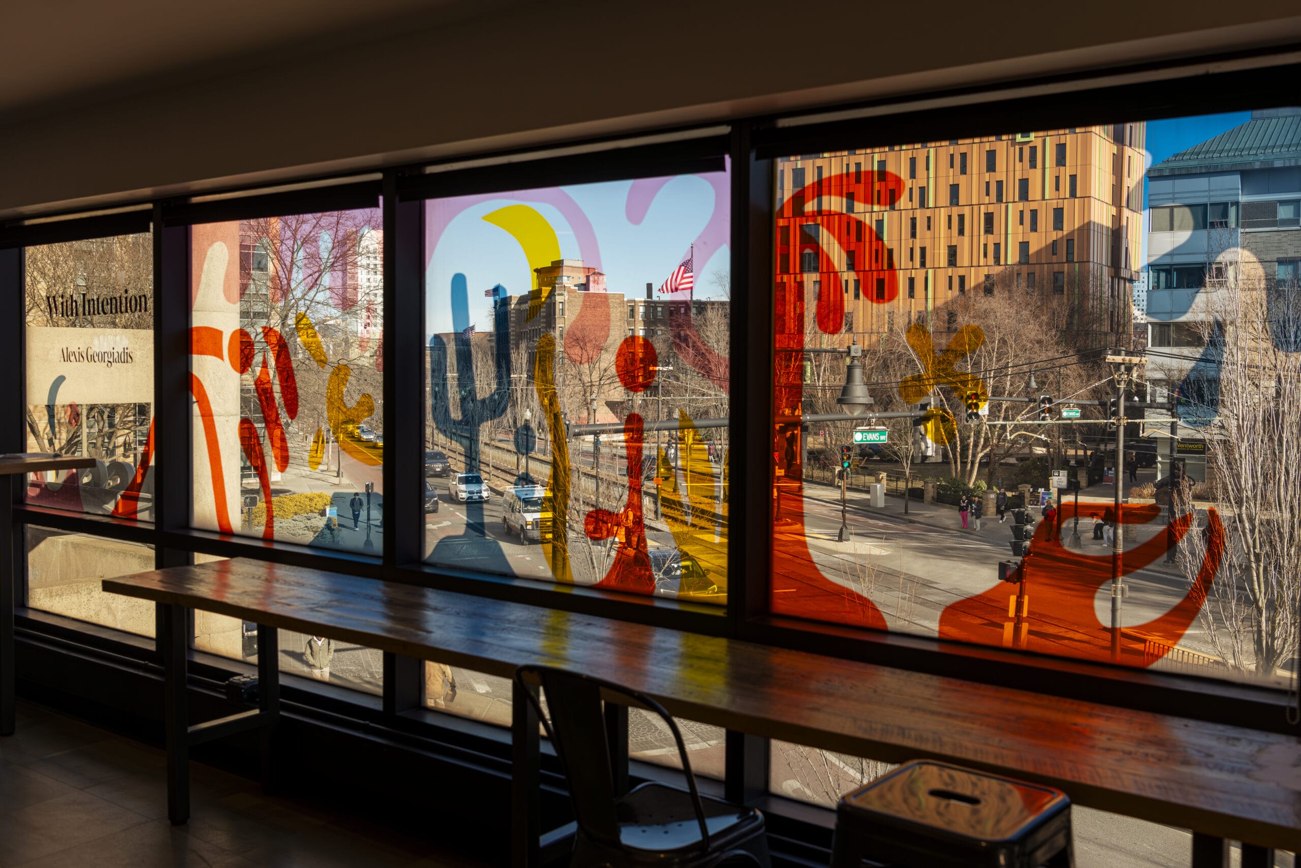 Colorful abstract shapes adorn large window panels overlooking a street scene with buildings and trees. A wooden table with bar stools stands against the window, suggesting a workspace that doubles as an informal Academic Resource Center, offering educational assistance to those who gather there.
