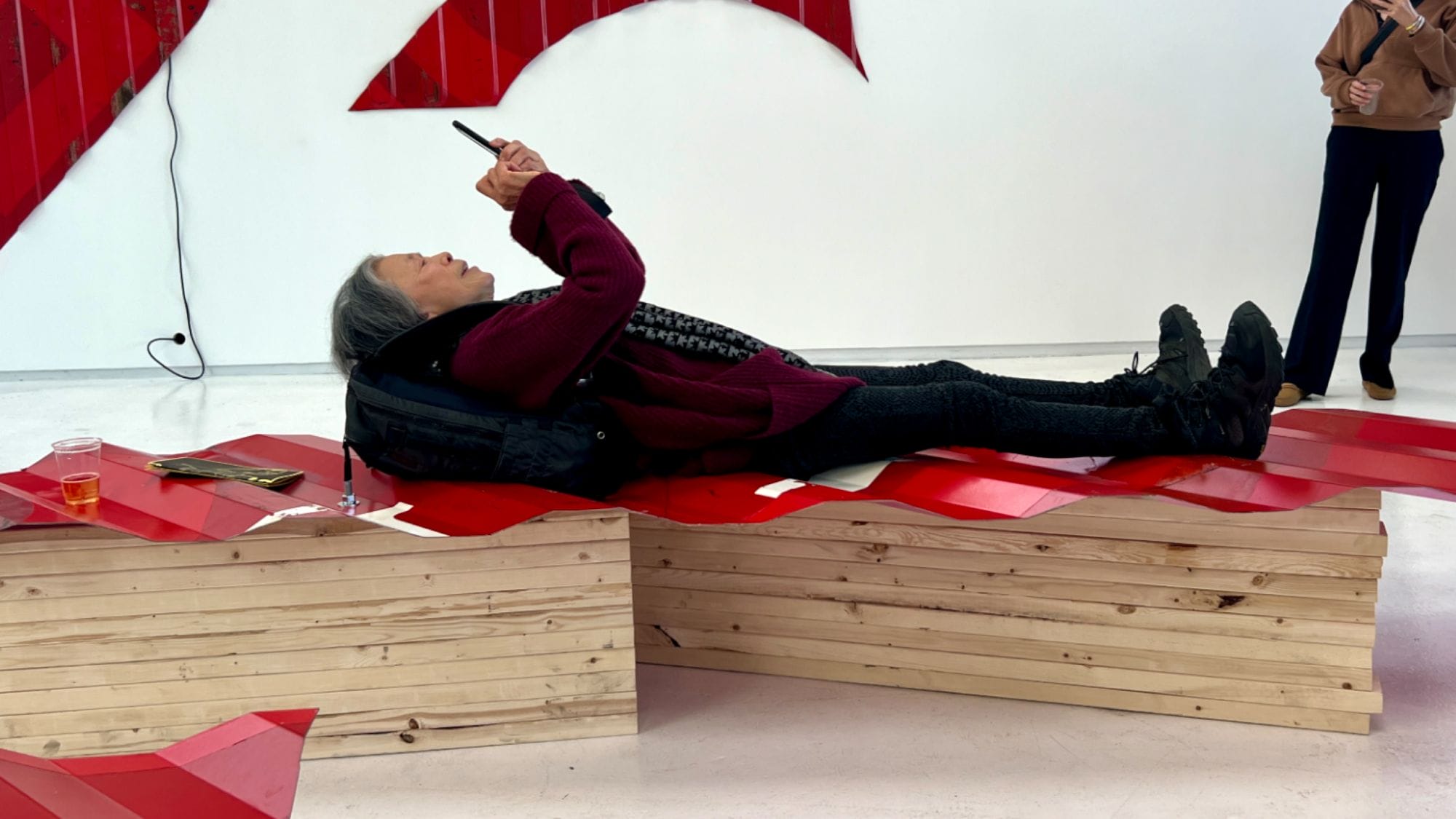 A person relaxes on a wooden bench featuring a red geometric design, absorbed in their phone. Clad in a dark jacket and boots, they encapsulate the essence of modern life. Nearby, MAAM-inspired abstract red wall art adds intrigue, while a drink rests beside them, completing this human tableau.