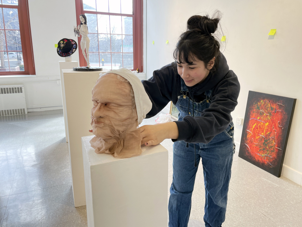 In an art studio filled with creations on pedestals and walls, a member of the student government with dark hair in a bun works diligently, wearing a hoodie and overalls while sculpting a head figure. Large windows flood the space with natural light, inspiring artistic expression.