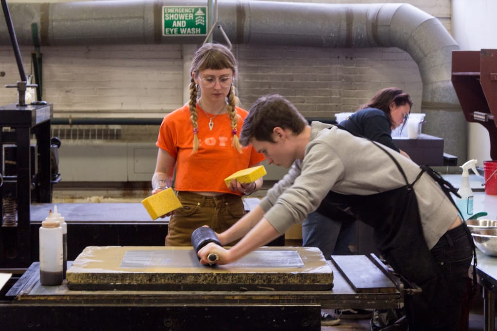 Students using a rolling pint to roll over prints in the printmaking studio