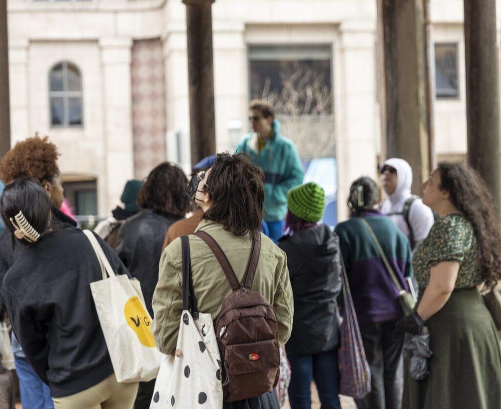 A group of people gathered outdoors, focusing on a speaker at the front. Several people wear jackets and backpacks. The setting appears to be urban, with a building facade in the background.