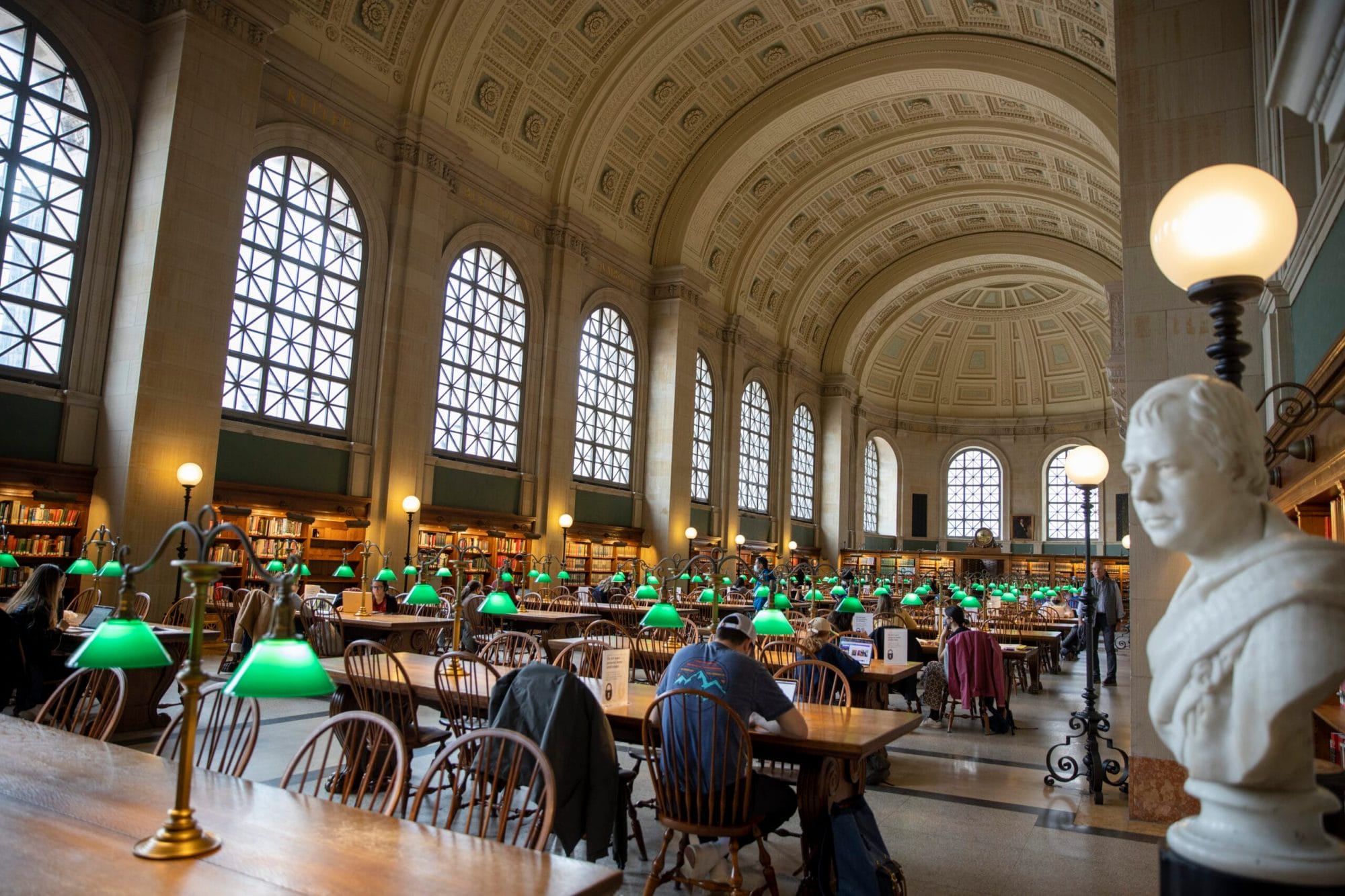 A grand library reading room with tall arched windows, ornate ceiling, and rows of wooden tables with green desk lamps. People are seated, reading and working. A white bust is prominently displayed in the foreground.