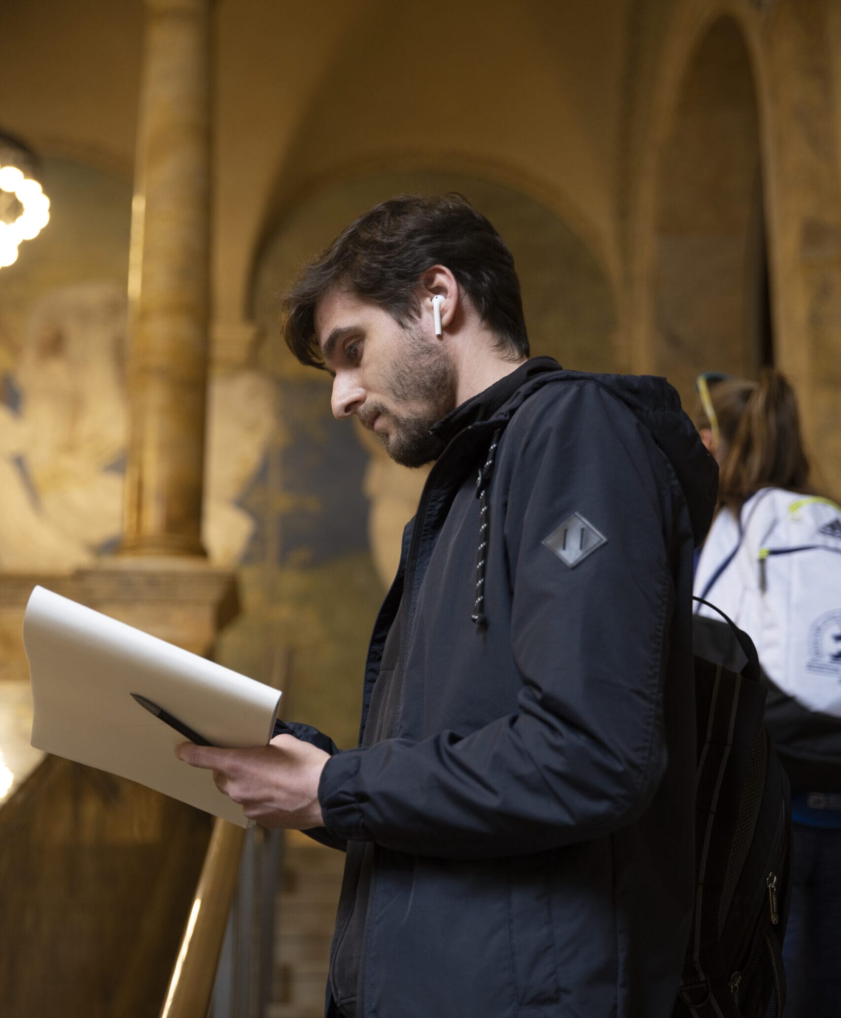 A man with earbuds stands indoors, holding a notebook and pen. He is dressed in a dark jacket, with soft lighting and architectural elements like pillars and arches in the background.