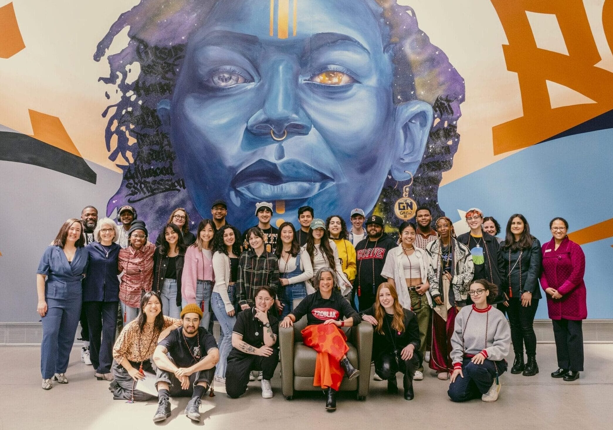 A group of MassArt staff from the Justice, Equity, and Transformation office gather with President Grant in front of a mural of a woman's face in the Design and Media Center.