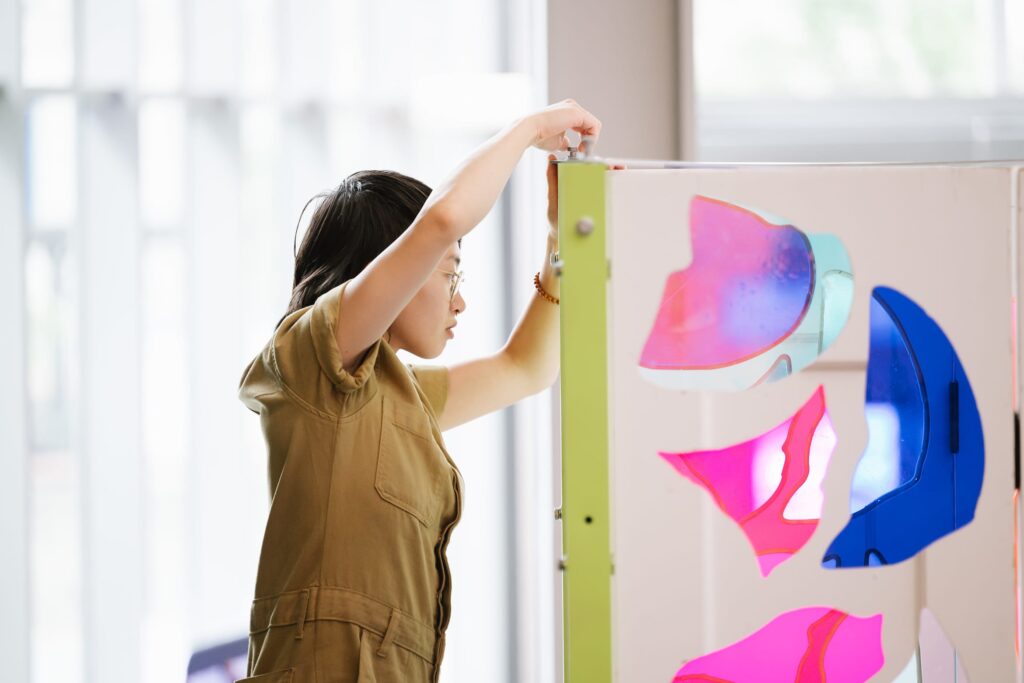 A woman holds the edge of a white, purple and pink artwork being installed at MassArt in the Design and Media Center.