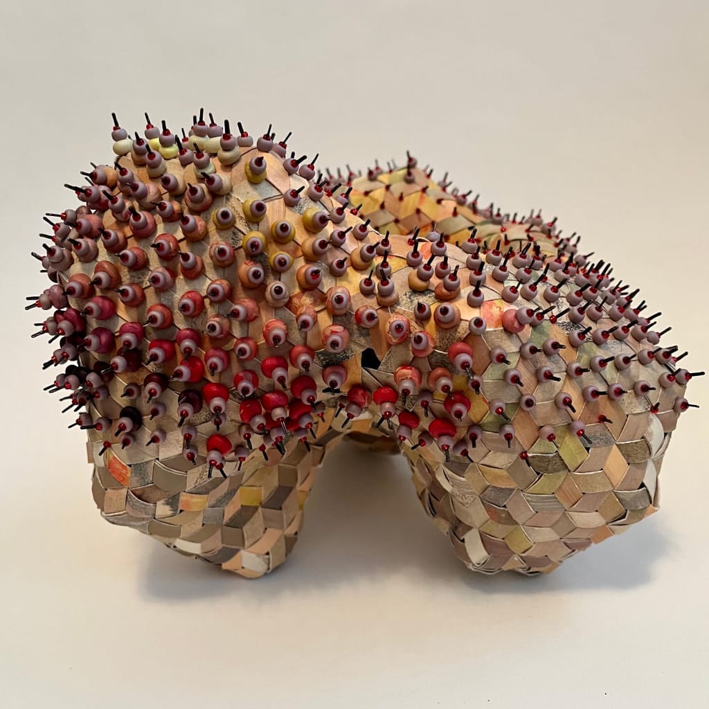 A sculptural artwork featuring two rounded forms covered in a mosaic of pencil stubs with sharpened tips pointing outward. The pencils vary in colors, creating a textured and spiky appearance against a neutral background.