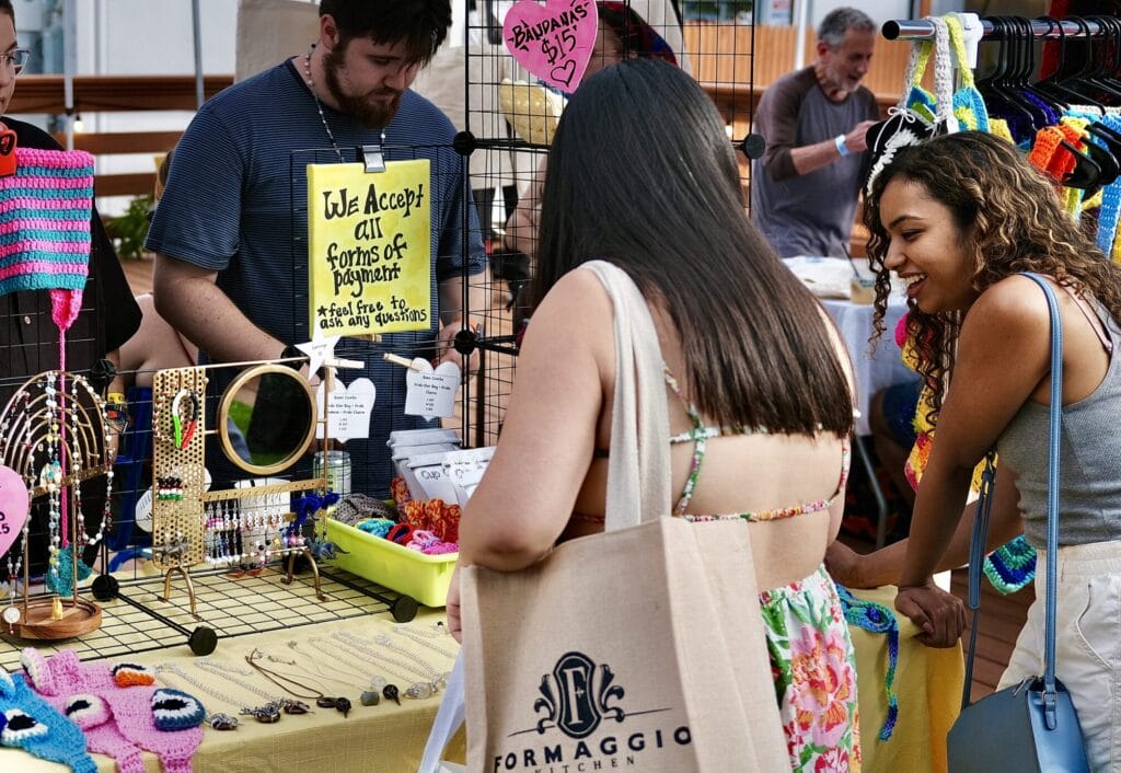 People browsing a colorful craft stall at the night market. The stall features handmade jewelry, crochet items, and a sign saying We Accept All Forms of Payment. One woman holds a reusable shopping bag, enjoying the art and chill vibe all around.