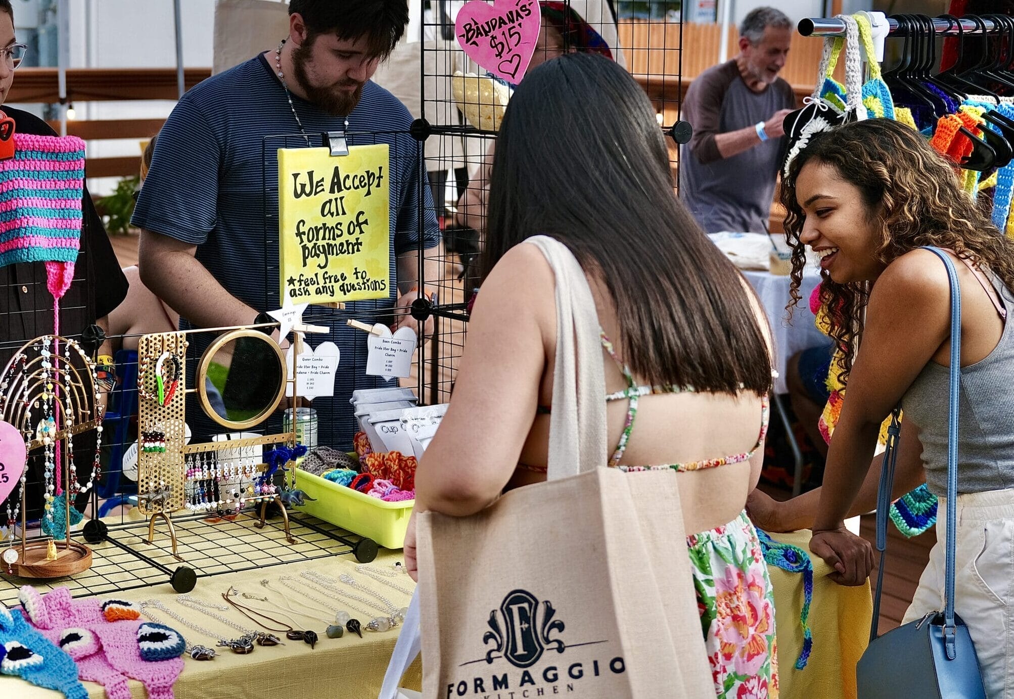 People browsing a colorful craft stall at the night market. The stall features handmade jewelry, crochet items, and a sign saying We Accept All Forms of Payment. One woman holds a reusable shopping bag, enjoying the art and chill vibe all around.